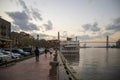 A red brick footpath along the Savannah River with the Talmadge Memorial Bridge over the water and ships docked along the banks