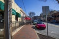 A red brick sidewalk with tables, chairs and red umbrellas with people walking and cars parked and cars driving on the street