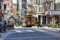 A red cable car riding up a street with restaurants and retails stores and people walking on the sidewalk at Union Square