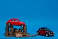 Red car figurine on top of a pile of coins next to a crimson car figurine, on blue background