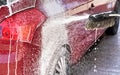 Red car washed in self serve carwash, detail on white soap foam spraying from brush / broom onto side above rear wheel