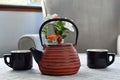 Red Cast Iron Teapot with black cups by gray chair near window ledge
