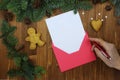 Red Christmas envelope with blank paper card, old woman`s hand with a pen. Fir branches, gingerbread man on a wooden background. Royalty Free Stock Photo