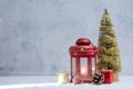 Red Christmas lantern glows beside a festive tree, ornaments, pinecone and gifts in a snowy winter setting. Holiday composition Royalty Free Stock Photo