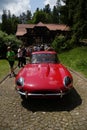 Red classic Jaguar E-Type parked on a cobblestone road surrounded by people