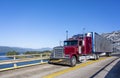 Red classic bonnet big rig semi truck transporting frozen cargo in bright refrigerated semi trailer driving on the truss bridge Royalty Free Stock Photo