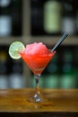 Red cocktail with ice garnished with lime standing on wooden bar counter. On background rack with bottles. Royalty Free Stock Photo