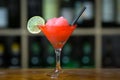 Red cocktail with ice garnished with lime standing on wooden bar counter. On background rack with bottles. Royalty Free Stock Photo