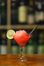 Red cocktail with ice garnished with lime standing on wooden bar counter. On background rack with bottles Royalty Free Stock Photo