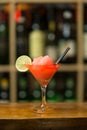 Red cocktail with ice garnished with lime standing on wooden bar counter. On background rack with bottles Royalty Free Stock Photo