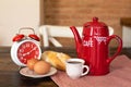 A coffee pot, a red alarm clock, a white cup of coffee and breakfast are on the wooden table. Royalty Free Stock Photo