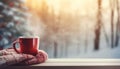 Red cup of coffee wrapped in wool scarf on table against winter forest background with blurred Christmas tree