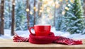 Red cup of coffee wrapped in wool scarf on wooden table against winter forest background with blurred Christmas tree