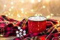 Red cup with hot coffee and sweet marshmallows on wooden table with plaid in Christmas decorations with garland lights