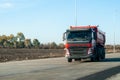 A red dump truck drives out of town on a flat asphalt road among fields against a blue sky on a sunny day Royalty Free Stock Photo