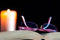 Red eyeglass on opened book, candle light.