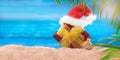 Red gift box with yellow bow and Santa Claus` hat on the beach behind sea and palm trees. Concept of Christmas, New year holiday Royalty Free Stock Photo