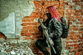 Red-hair airsoft woman in uniform with machine-gun, stand beside brick wall inside broken building