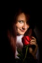 A red-haired beautiful girl with a rose in her hand smiles a malicious smile on a black background.