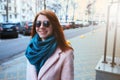 Red haired beautiful girl is walking by the street in a pink coat and blue scarf, with sunglasses. Royalty Free Stock Photo