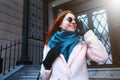 Red haired beautiful girl is walking by the street in a pink coat and blue scarf, with sunglasses. Royalty Free Stock Photo