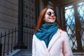 Red haired beautiful girl is walking by the street in a pink coat and blue scarf, with sunglasses. Royalty Free Stock Photo