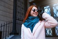 Red haired beautiful girl is walking by the street in a pink coat and blue scarf, with sunglasses. Royalty Free Stock Photo