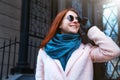 Red haired beautiful girl is walking by the street in a pink coat and blue scarf, with sunglasses. Royalty Free Stock Photo