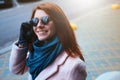 Red haired beautiful girl is walking by the street in a pink coat and blue scarf, with sunglasses. Royalty Free Stock Photo