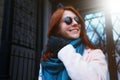 Red haired beautiful girl is walking by the street in a pink coat and blue scarf, with sunglasses. Royalty Free Stock Photo