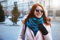 Red haired beautiful girl is walking in the urban background in a pink coat and blue scarf, with sunglasses. Royalty Free Stock Photo