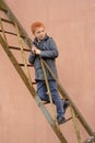 Red-haired boy  in a grey coat climbed the rusty metal ladder to the wall and looks back suspiciously Royalty Free Stock Photo