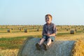 A red-haired boy sits on top of a straw bale on a wheat field