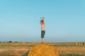 A red-haired boy stands with hands up on top of a straw bale on a wheat field