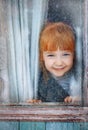 Red-haired charming girl looks out the window and smiles. Winter snow-covered window and blue-eyed girl looking at the camera Royalty Free Stock Photo