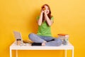 Red haired female wearing green t shirt and jeans sitting on table with crossed legs, being tired from learning, making spyglass