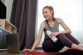 A red-haired girl with a bottle in her hand looks at her laptop with a smile, sitting on the floor in a sports uniform.