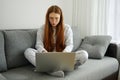 A red-haired girl with glasses, with long hair in pajamas sits cross-legged on the couch and looks intensely at the Royalty Free Stock Photo