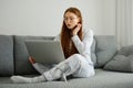 A red-haired girl with glasses, with long hair in pajamas sits cross-legged on the couch and looks at the laptop Royalty Free Stock Photo