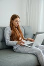 A red-haired girl with glasses, with long hair in pajamas sits on the sofa in a half-turn and looks at the laptop Royalty Free Stock Photo