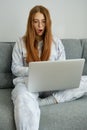 A red-haired girl with glasses, with long hair sits on the couch in a half-turn and looks in surprise at the laptop Royalty Free Stock Photo