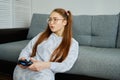 A red-haired girl with glasses, with long tails, sits on the floor in a half-turn leaning on a sofa with a game console Royalty Free Stock Photo