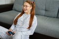 A red-haired girl with glasses, with long tails sits on the floor, leaning on a sofa and joyfully plays on a game Royalty Free Stock Photo