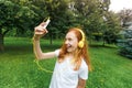 Red-haired girl listens to lessons in yellow headphones. Teenager in white t-shirt and headphones in the park