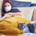 Red-haired girl in a medical mask and a cat lie on the bed Royalty Free Stock Photo