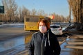 A red-haired girl in a medical mask stands at a public transport stop on a sunny spring evening.