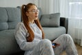 A red-haired girl in pajamas and glasses sits by the sofa with a game joystick and looks with enthusiasm in front of her Royalty Free Stock Photo
