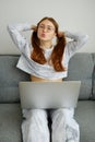 A red-haired girl in pajamas and glasses sits on a sofa with a laptop, collecting her hair in ponytails and stretching Royalty Free Stock Photo