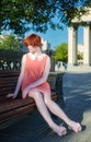 Red haired girl in a short pink dress on a park bench