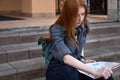 Red-haired girl sits on the steps with a backpack and looks at the map thoughtfully, the wind blows her hair Royalty Free Stock Photo